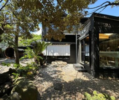 Gravel courtyard with stone pathways and trees beside a dark wooden ryokan building with glass windows