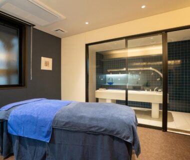 Massage table with blue linens and glass-partitioned wash area at AMANE resort GAHAMA in Oita, Kyushu