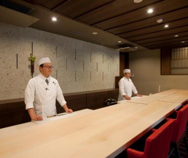 Two chefs behind a long wooden counter with red chairs at AMANE resort GAHAMA, Ōita, Kyūshū, Japan