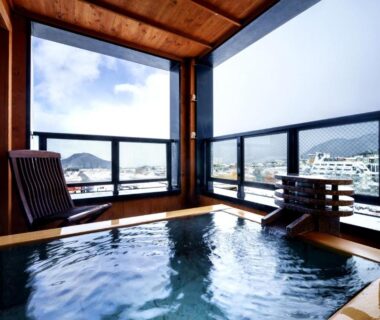 Wooden indoor onsen bath with balcony seating overlooking Nagano town and surrounding mountains at Aburaya Tousen