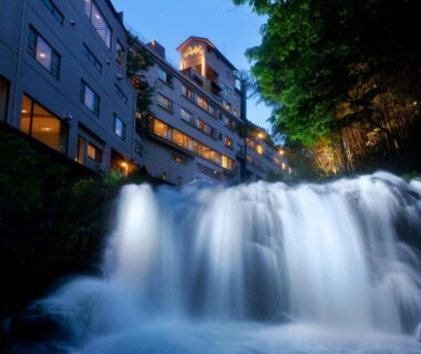 Cascading waterfall before illuminated Shosuke no Yado Takinoyu building on riverside in Fukushima