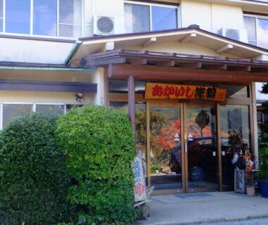 Entrance of Akaishi Ryokan with glass doors, hanging sign, potted plants and trimmed shrubs in Yamanashi