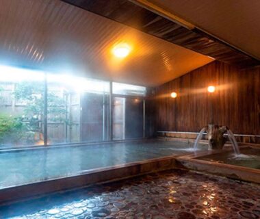 Indoor communal onsen with wooden paneling, stone floor and garden view through large glass window