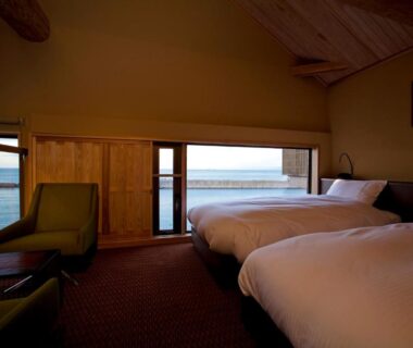Twin beds with lounge chair, wooden interior and sea visible through low windows
