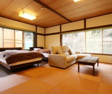 Tatami-floored room with twin beds, sofa, low table and large windows showing garden greenery at Amis Droles Shizuoka