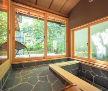 Indoor stone-tiled soaking bath with wooden tub and garden-view windows at Arai Ryokan, Shizuoka