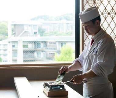 Uniformed chef arranging table service by window with wooden interior and town view at Arima Onsen ryokan