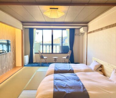 Tatami room with two twin beds, low table, chairs by large window and wall-mounted television at Ashiwada Hotel Yamanashi