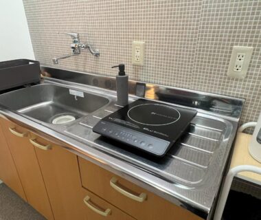 Compact stainless kitchenette with portable induction hob, electric kettle and cabinets at Assi House in Ōita, Kyūshū