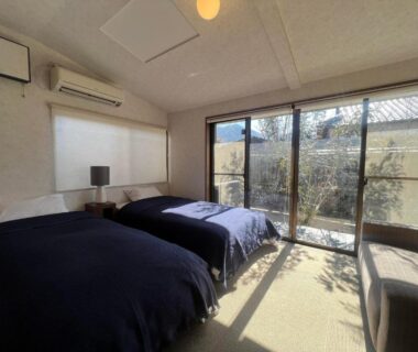 Bright tatami room with two beds, low table and sliding glass doors overlooking a courtyard garden at Assi House, Oita