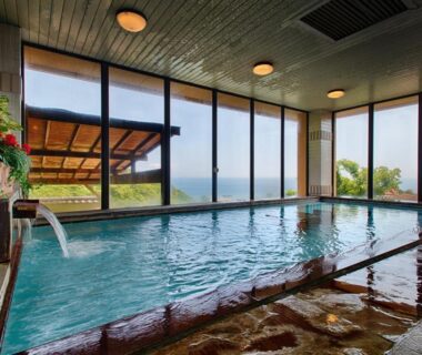 Spacious indoor onsen pool with stone edging, wooden ceiling and large windows with water beyond