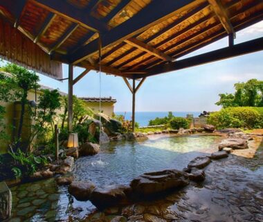 Covered stone onsen bath with wooden roof, garden plants and distant coastal view at Atagawa Prince Hotel Shizuoka