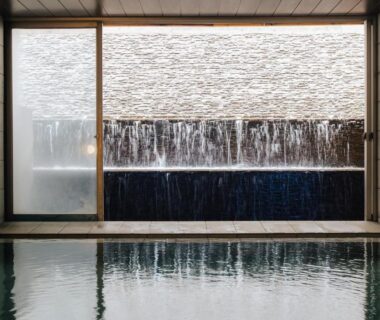 Indoor onsen pool with framed cascading water wall and stone tiles at Atami Fuga, Shizuoka