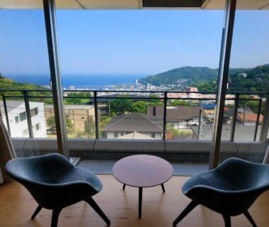 Two chairs and small table on balcony overlooking coastal town and distant sea from Atami Fuga room