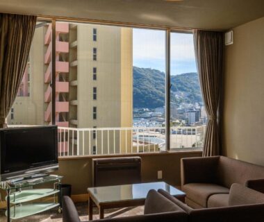 Sofa seating and table in lounge opening to balcony with city and hillside views at Atami Onsen Sakuraya Ryokan, Shizuoka