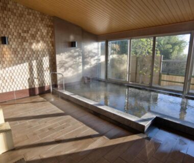 Spacious indoor onsen with wooden floor, tiled wall, stools, and floor-to-ceiling windows overlooking greenery