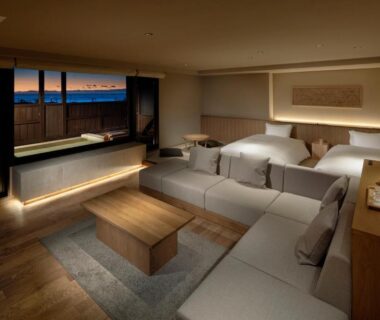 Spacious modern guest room with twin beds, sofa, low wooden table, in room wooden bath by large window at dusk