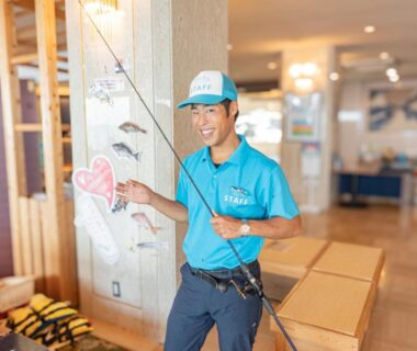 Smiling ryokan staff in blue uniform holding a fishing rod in a wooden lobby at Awakan Hyōgo