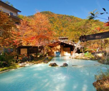 Outdoor onsen with wooden bathhouses surrounded by autumn maple foliage in Nagano mountains