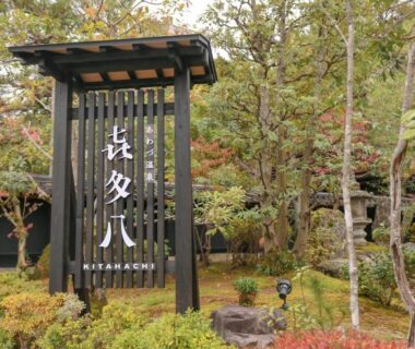 Wooden vertical entrance sign with kanji, surrounded by landscaped shrubs and trees at Awazuonsen Kitahachi, Ishikawa
