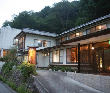 Evening exterior of Ayameya Ryokan with lit windows, wood entrance and hillside setting in Gunma