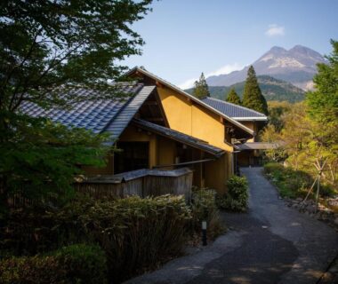 Wooden ryokan buildings along a quiet path with distant mountain and surrounding greenery in Ōita