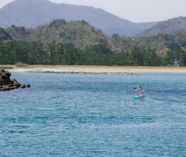 Paddleboarder near sandy shore with forested hills, view associated with Base House Kinosaki in Hyōgo, Kansai