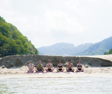 Group seated on sandy riverside near mountains at Base House Kinosaki, Hyōgo, Japan