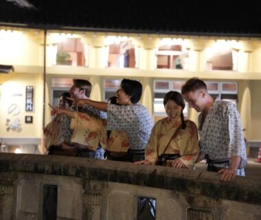 Group in yukata standing on a stone bridge at night with illuminated town buildings behind