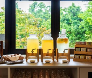Three glass beverage dispensers and cups on wooden counter by large window with green trees at Beppu Fuga, Ōita