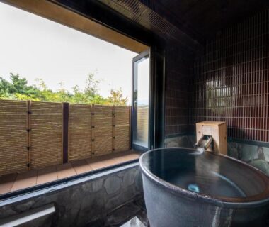 Stone onsen tub beside tiled wall with open sliding door to bamboo-fenced balcony and green garden beyond