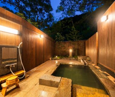 Wood-lined open-air onsen bath with stone edging, shower fixtures and wooden stool at Beppu Showaen Oita