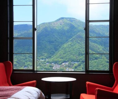 Window-framed mountain view from guestroom with red armchairs, round table and bed at Bettei Koyoi in Kanagawa
