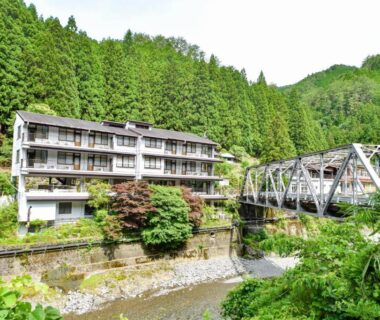 Three-story ryokan beside river with metal truss bridge and dense evergreen hills in Wakayama, Kansai