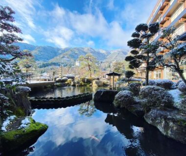 Reflective garden pond with trimmed pines, light snow on rocks, mountain backdrop at Bosenkan in Chūbu, Gifu, Japan