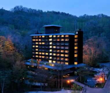 Illuminated multi-story ryokan building at dusk surrounded by wooded hills in Noboribetsu, Hokkaidō