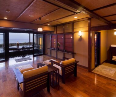 Spacious sitting area with wooden floors, low table and balcony doors opening to calm coastal waters at Bousui, Shizuoka