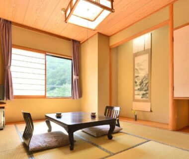 Japanese tatami room with low table, floor chairs, alcove with hanging scroll, TV and window at Bouzantei Kotobuki, Nagano