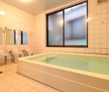 Tiled communal bathing room with rectangular soaking tub, stools, faucets and frosted window at Business Ryokan Iroha in Gifu