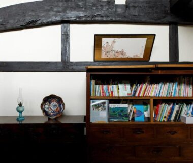 Interior bookshelf with books, decorative plate and lamp beneath exposed wooden beams at Busuitei ryokan