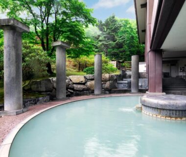 Outdoor hot spring pool with pale milky water and stone pillars amid green trees at Dai-ichi Takimotokan, Hokkaidō