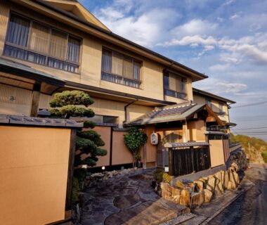 Two-story traditional ryokan exterior with wooden balconies, stone entrance path and coastal road in Hyōgo