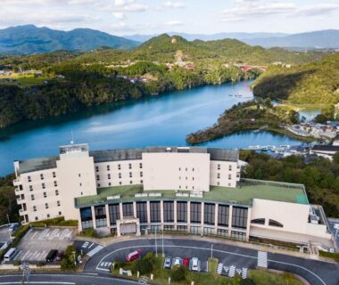 Curved hotel building along a calm river with surrounding forested hills and winding shoreline