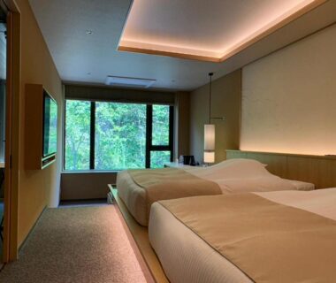 Minimal twin beds and wooden interior with large window at Exclamation Hotel in Hokkaidō showing leafy forest view