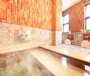 Steaming indoor onsen bath with marble lower walls, wooden upper cladding, steps into water and traditional shower stools