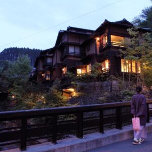 Guests walking on wooden bridge past illuminated riverside ryokan at dusk in Kumamoto