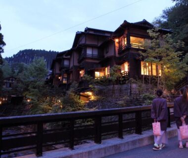 Guests walking on wooden bridge past illuminated riverside ryokan at dusk in Kumamoto
