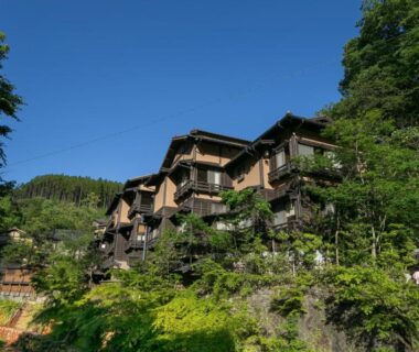 Multi-story traditional wooden inn with balconies among trees on a sunlit hillside in Kumamoto, Kyushu