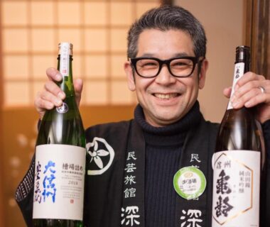 Smiling ryokan staff holding two sake bottles with shoji screens in Fukashiso, Nagano