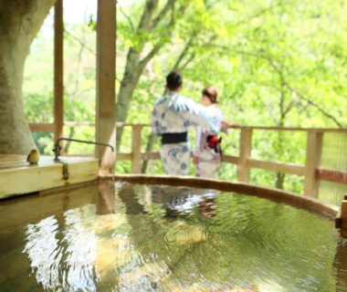 Outdoor wooden onsen tub with two guests in yukata overlooking green forest at Fukeikan 風景館 Nagano Chūbu Japan
